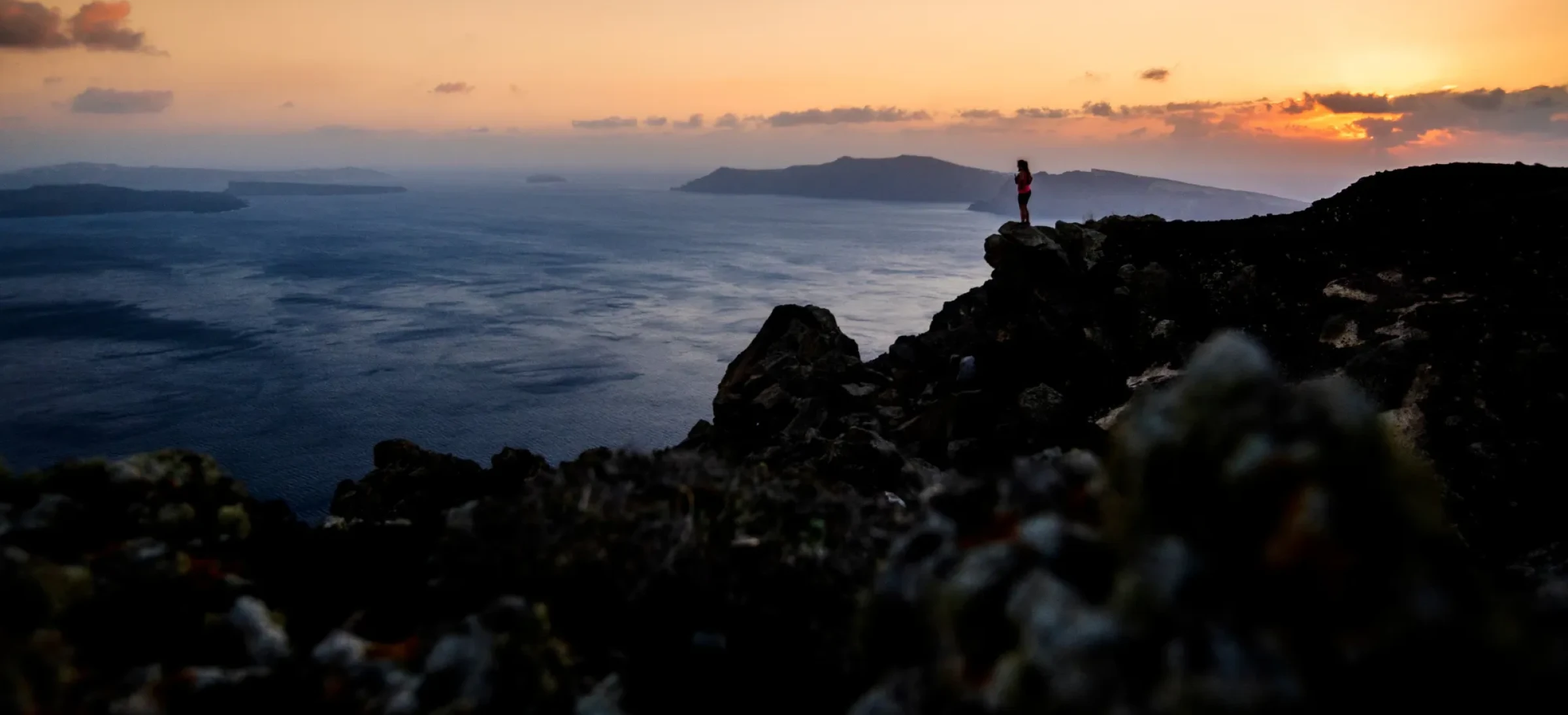 Person stood on rocky ledge, looking out at sunset
