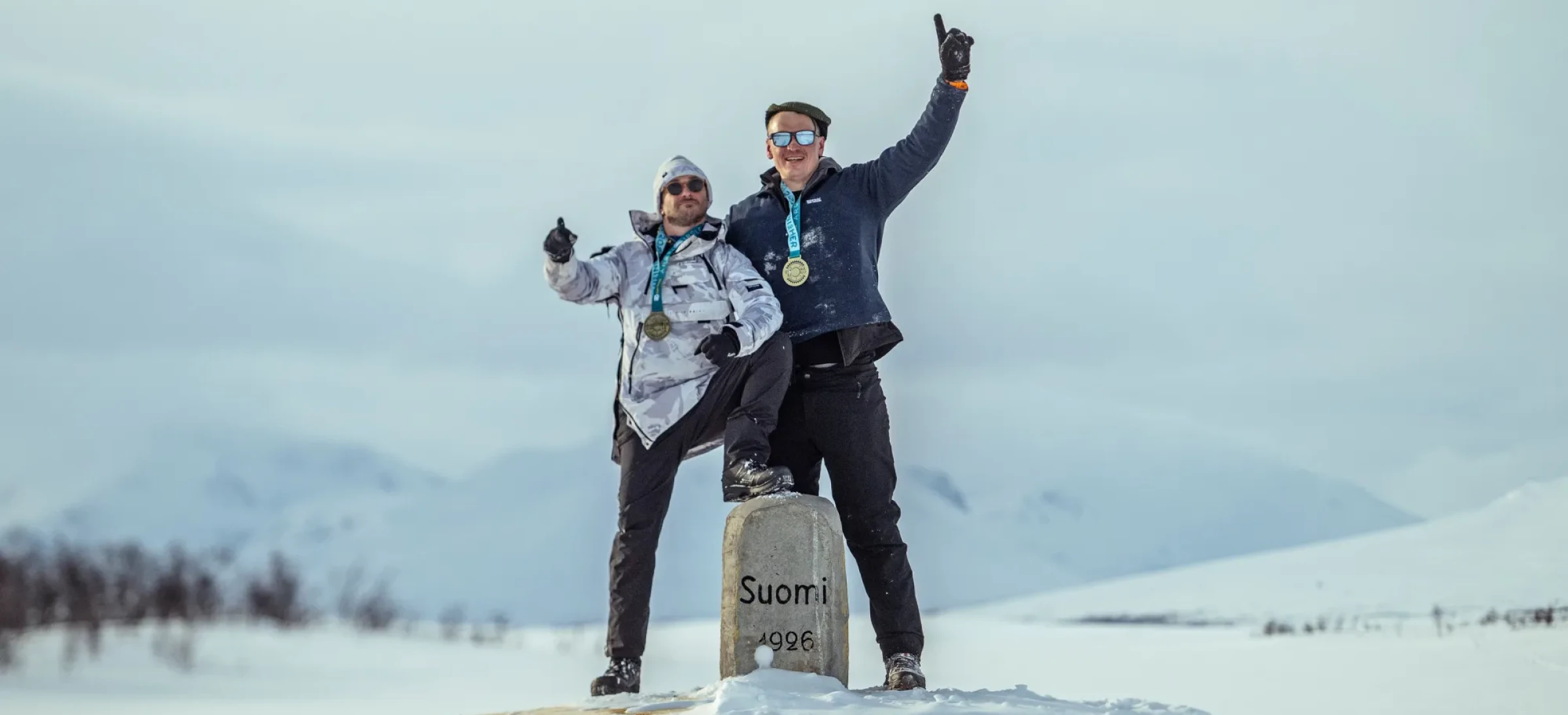 Two participants wearing medals standing on a snow-covered yellow marker in a winter mountain landscape, celebrating an achievement in snowy conditions.