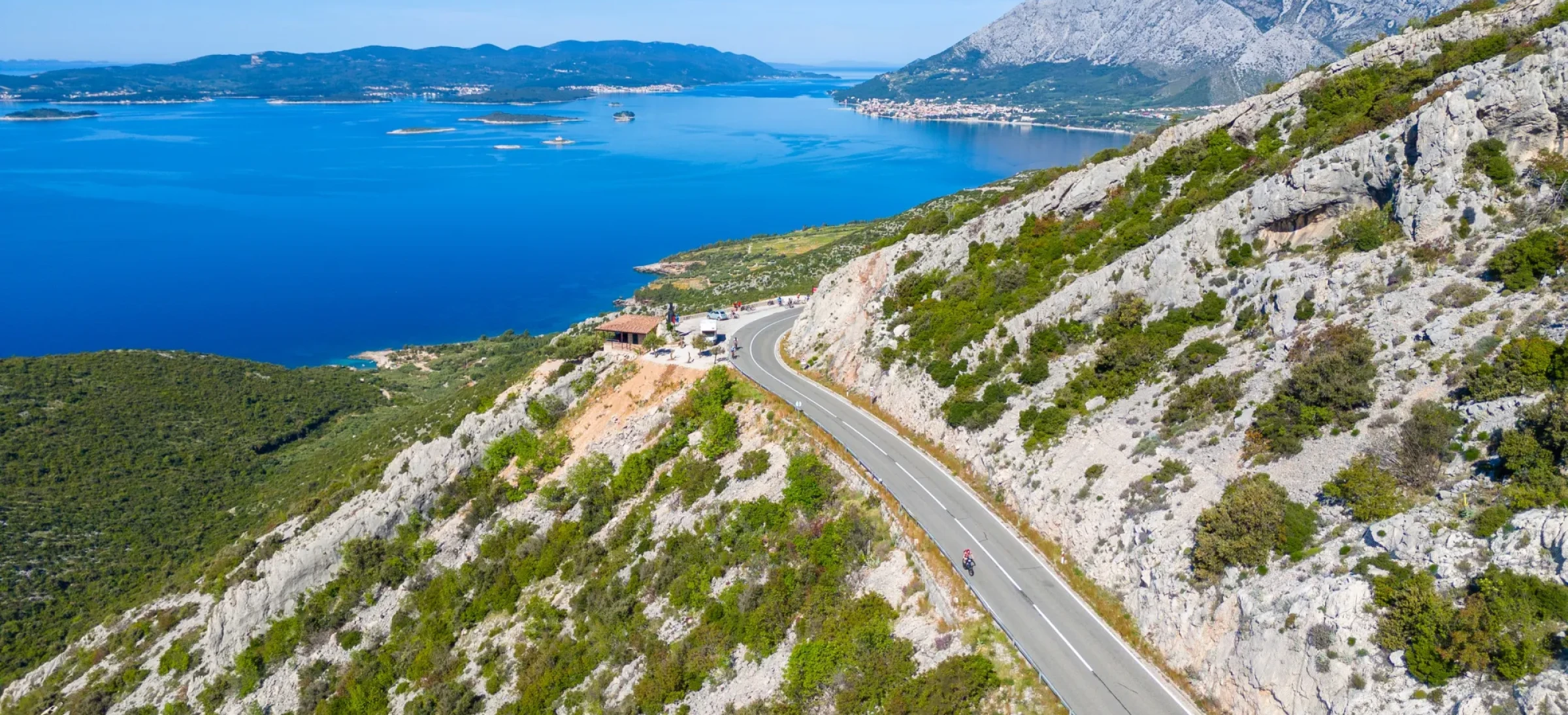 Cyclists ride along a road carved through a cliffside, with the bright blue sea visible in the background