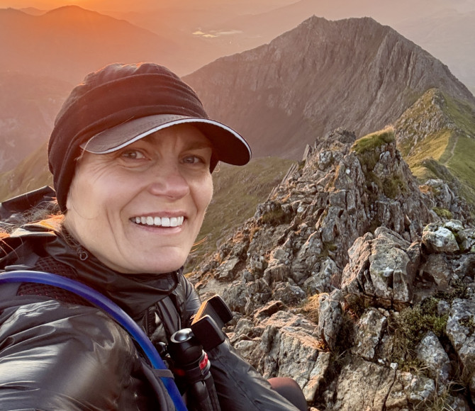Woman sat on a mountain with the sunset behind her and smiling