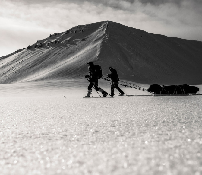 Dark image of participants pulling sleds through snow with a mountain behind