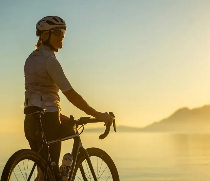 A blonde female cyclist looks to her right, with sunlit water and mountains stretching across the background