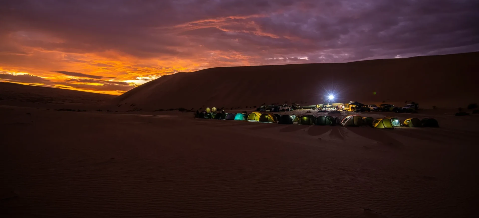 Tents next to sad dunes with dark, colourful sky behind