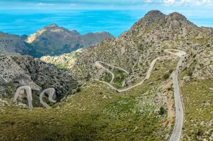 A winding road snakes through rugged, rocky terrain dotted with patches of greenery beneath a bright blue sky