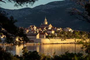 Buildings enclosed by a stone wall sit beneath mountainous scenery, with water stretching around