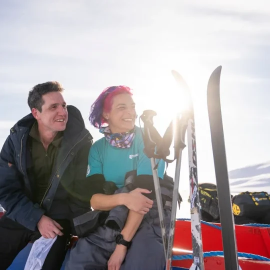 Woman with colourful hair and man sitting beside skis in a snowy mountain setting and both smiling