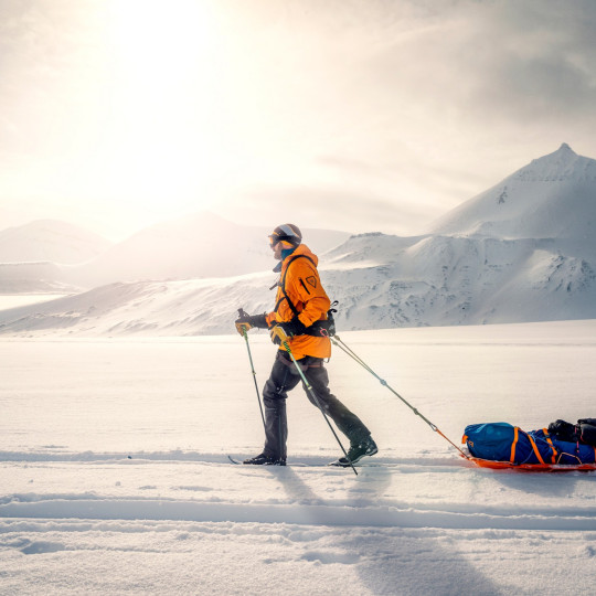 Participant pulling a sled through snow and snow-covered mountains in background