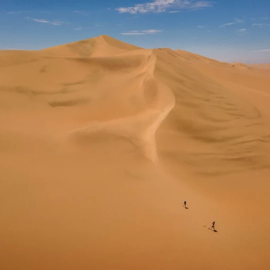 Two people running down smooth sand dunes with bright sky above