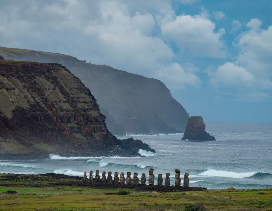 Statues stood infront of crashing waves and a mountainous terrain, with blue sky and clouds behind