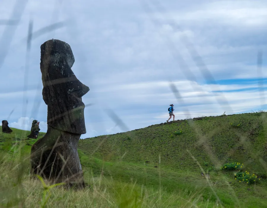 A large statue and woman running over a grassy hill with blue sky above her