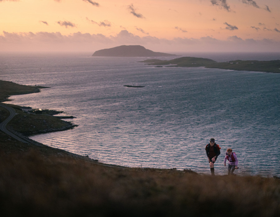 Two people walking up hill with water and colourful sky behind