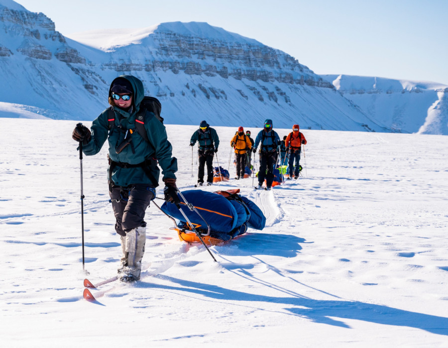Participants pulling sleds through snow with a bright sky above them and snow-covered mountains in the background