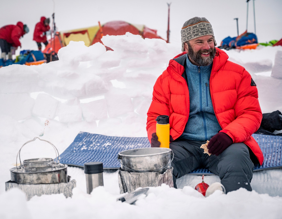 Man sat on snow, wearing a bright coloured coat and smiling