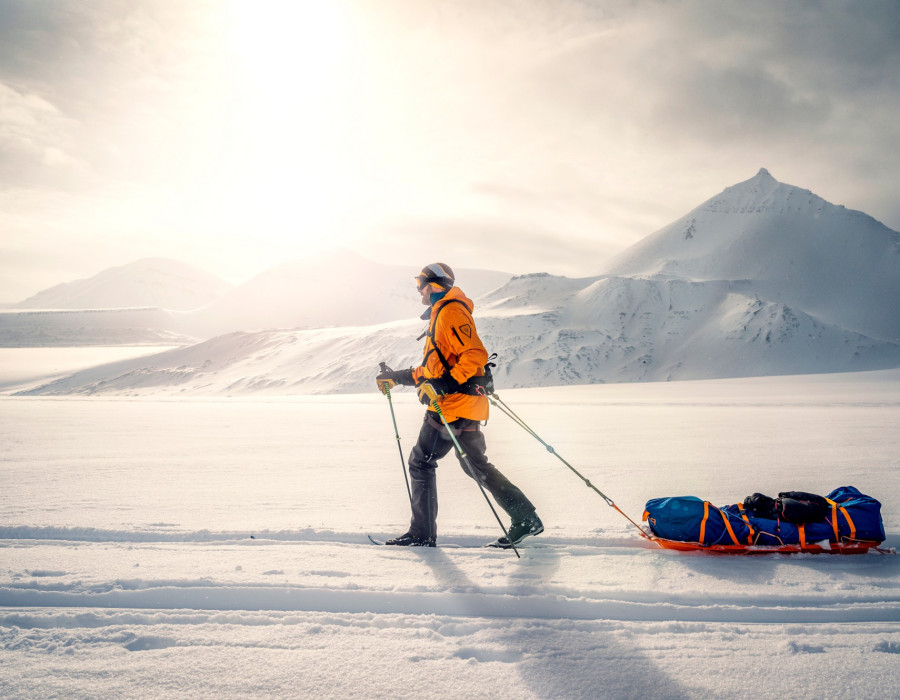 Participant pulling a sled through snow and snow-covered mountains in background