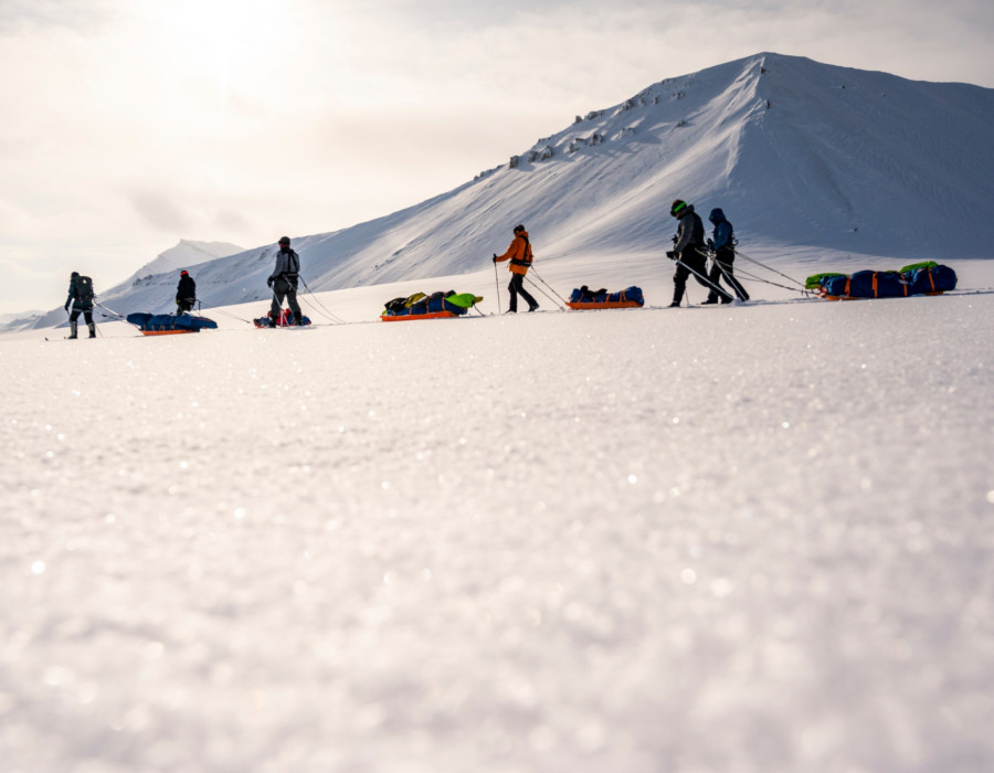 Participants pulling sleds through snow with a bright sky above them and snow-covered mountains in the background