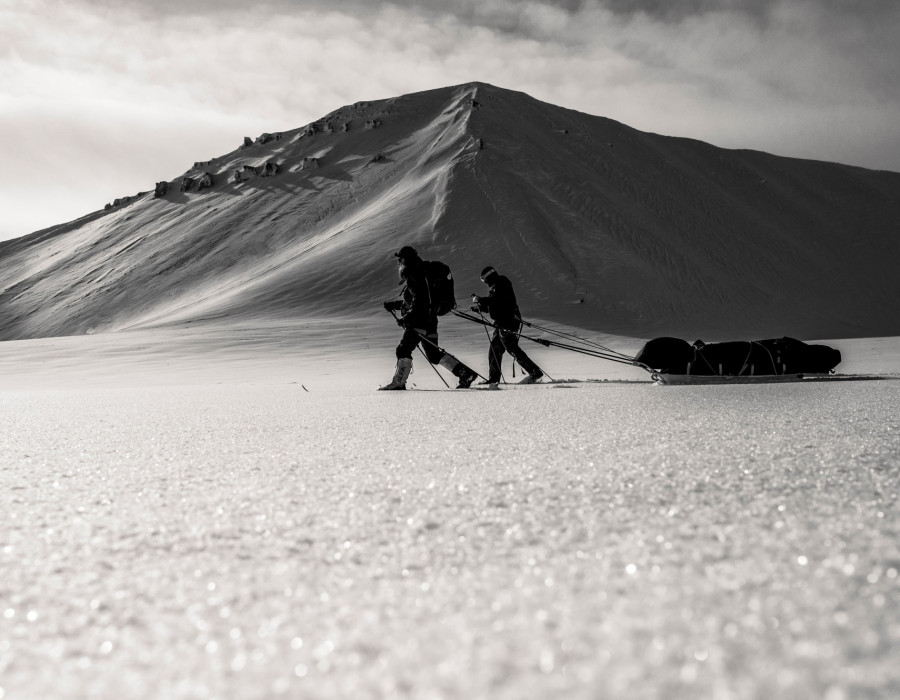 Dark image of participants pulling sleds through snow with a mountain behind
