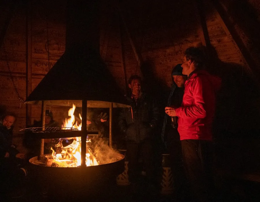 Group of participants gathered around a fire inside a wooden shelter, illuminated by warm firelight in a dark setting.