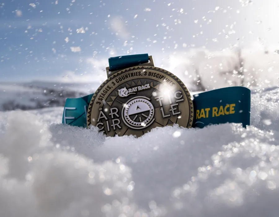 Close-up of an Arctic Circle race medal resting in the snow, with sunlight reflecting off the icy winter landscape.