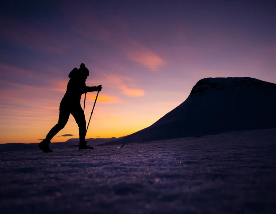 Silhouette of a lone participant crossing a snowy landscape at sunset, with a snow-covered mountain beneath a colourful twilight sky