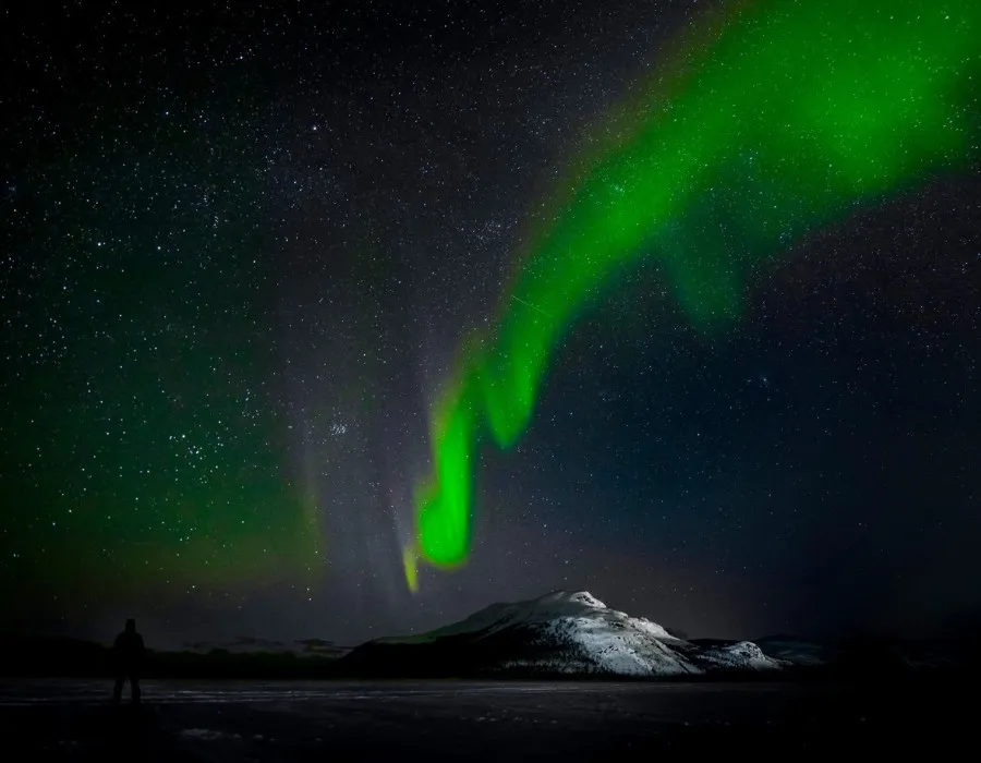 Man standing beneath a snowy mountain, looking up at a dark starry sky illuminated by bright green northern lights