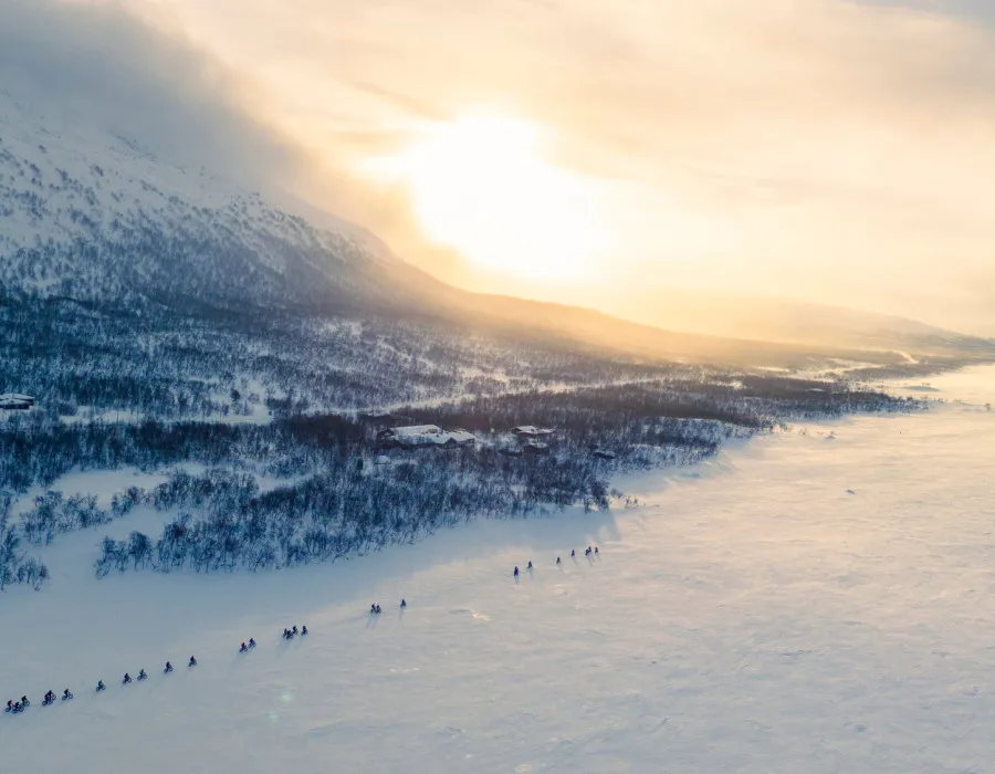 Aerial view of cyclists riding across a frozen snow-covered landscape at sunrise, surrounded by mountains and soft winter light.