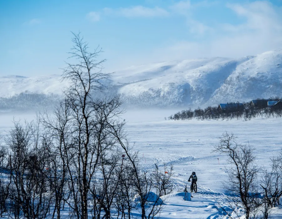 Single cyclist riding a fat bike across a snowy landscape, with mountains in the background and a blue sky above