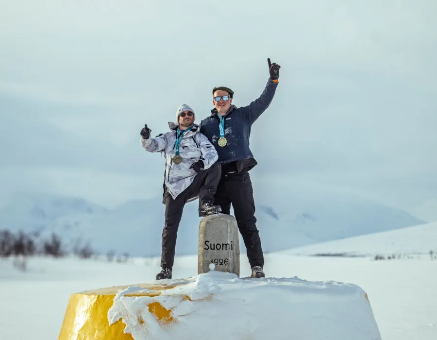 Two participants wearing medals standing on a snow-covered yellow marker in a winter mountain landscape, celebrating an achievement in snowy conditions.