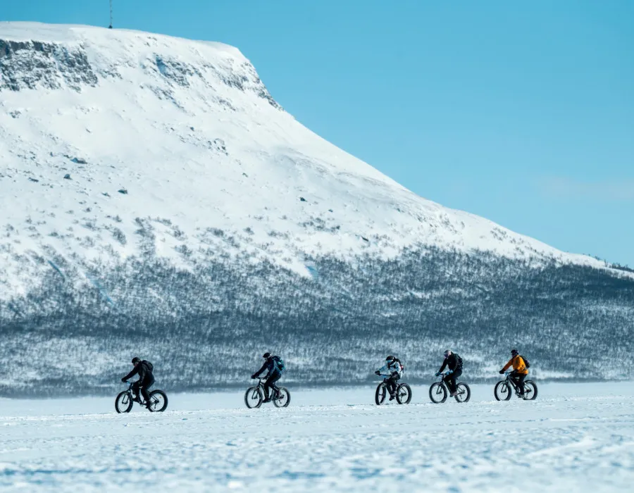 Group of cyclists riding fat bikes across a snow-covered landscape beneath a towering mountain and clear blue winter sky.