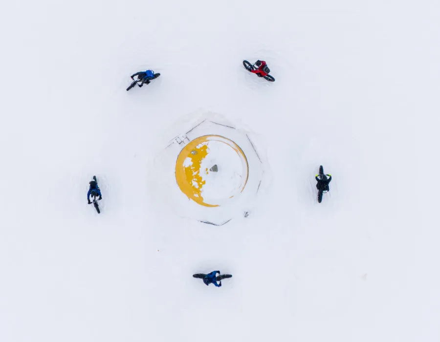 Aerial view of five cyclists riding fat bikes in a circle around a snow-covered yellow structure in a vast white winter landscape