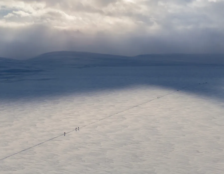 Aerial view of participants travelling across a vast snow-covered landscape beneath dramatic clouds and shifting winter light.