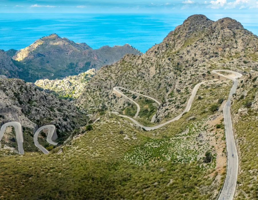 A winding road snakes through rugged, rocky terrain dotted with patches of greenery beneath a bright blue sky