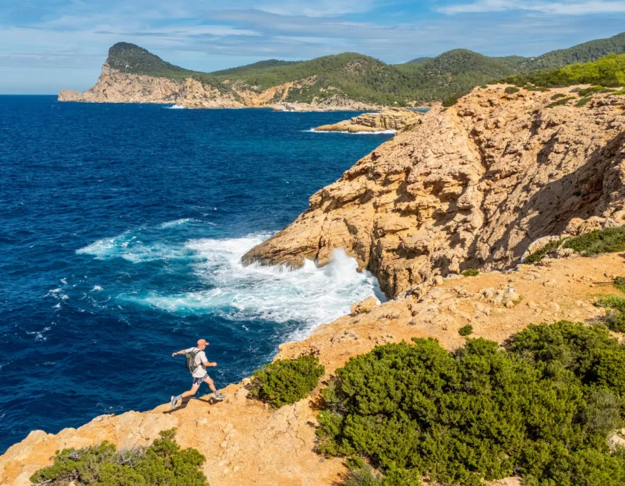 An event participant runs along the edge of a cliff beside a deep blue sea, with orange-red rock, patches of greenery, and a sunny sky above