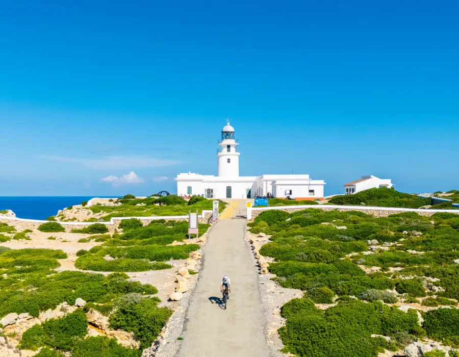 Cyclist riding away from a white lighthouse, with a bright blue sky above and patches of greenery at either side of the road