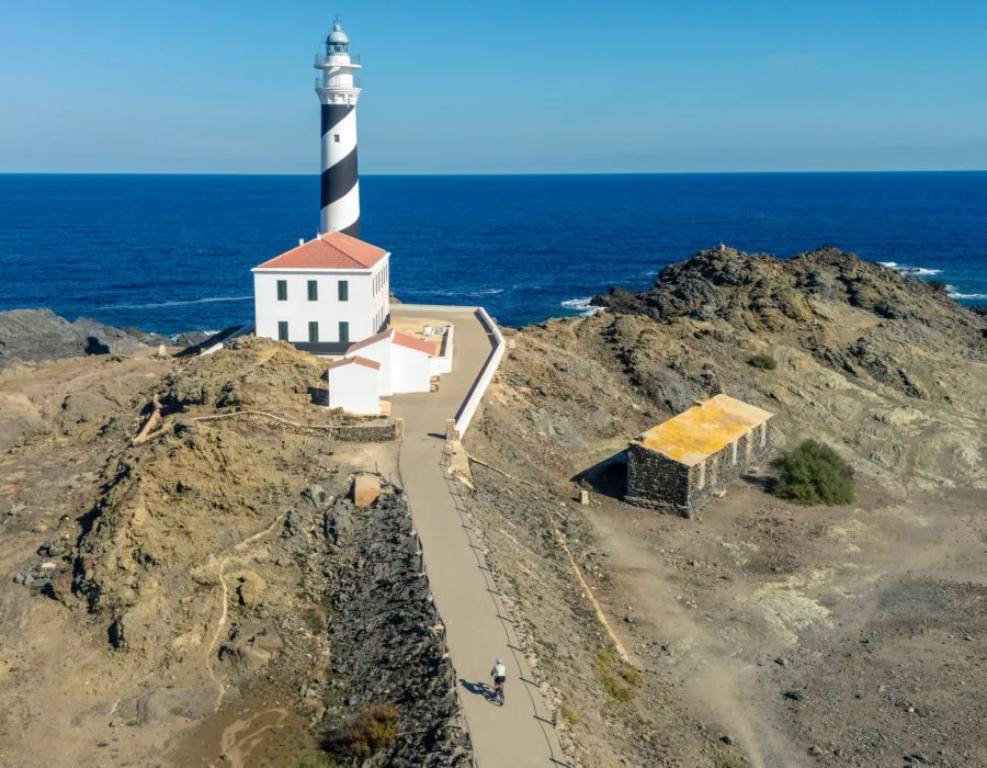 A cyclist rides towards a white lighthouse perched on the edge of a cliff overlooking the sea