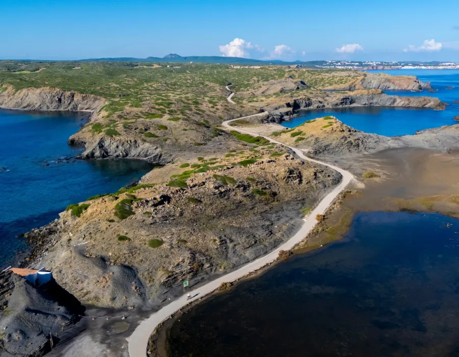 An aerial view of a winding coastal road cutting through a rugged rocky landscape, bordered by deep blue sea and lagoons under a clear blue sky