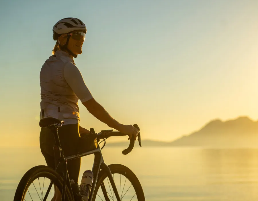 A blonde female cyclist looks to her right, with sunlit water and mountains stretching across the background
