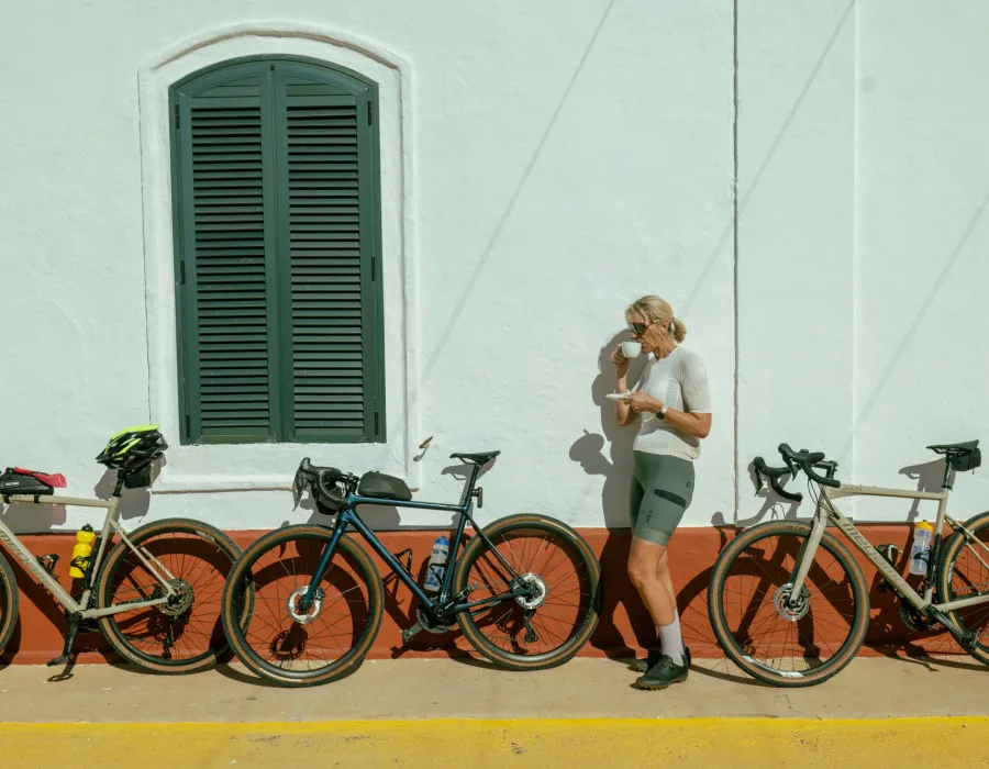 A blonde woman stands in front of a white wall, drinking from a mug, with three bicycles leaning against the wall behind her