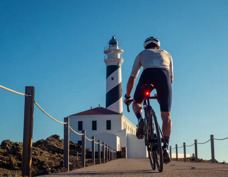 A cyclist riding along a path towards a lighthouse and a clear blue sky above
