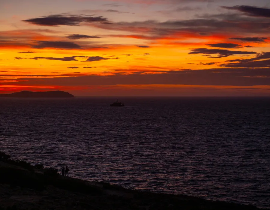 Two silhouettes look out onto a dark sea, with an orange and red sky above