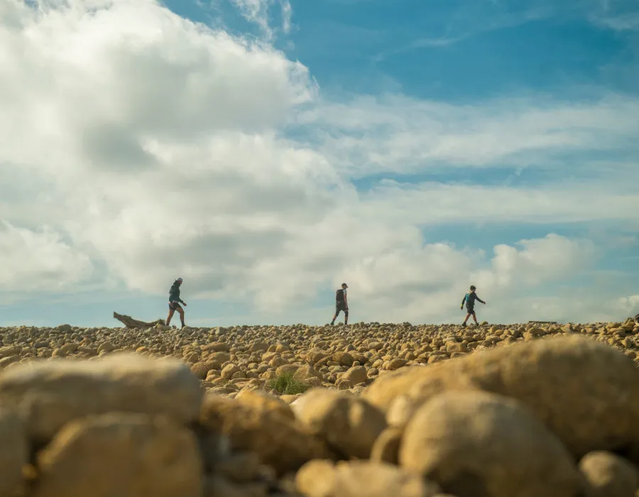 Three event participants walk across pebbly, sand-coloured ground beneath a blue sky with clouds.