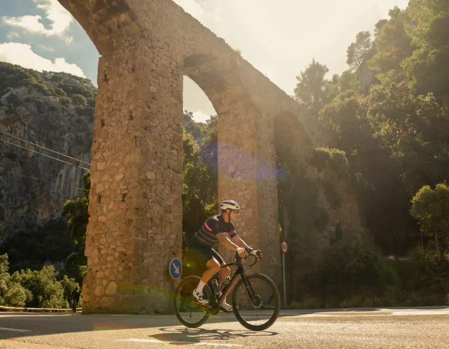 A lone cyclist rides beneath a viaduct, surrounded by trees with a rugged rockface behind them