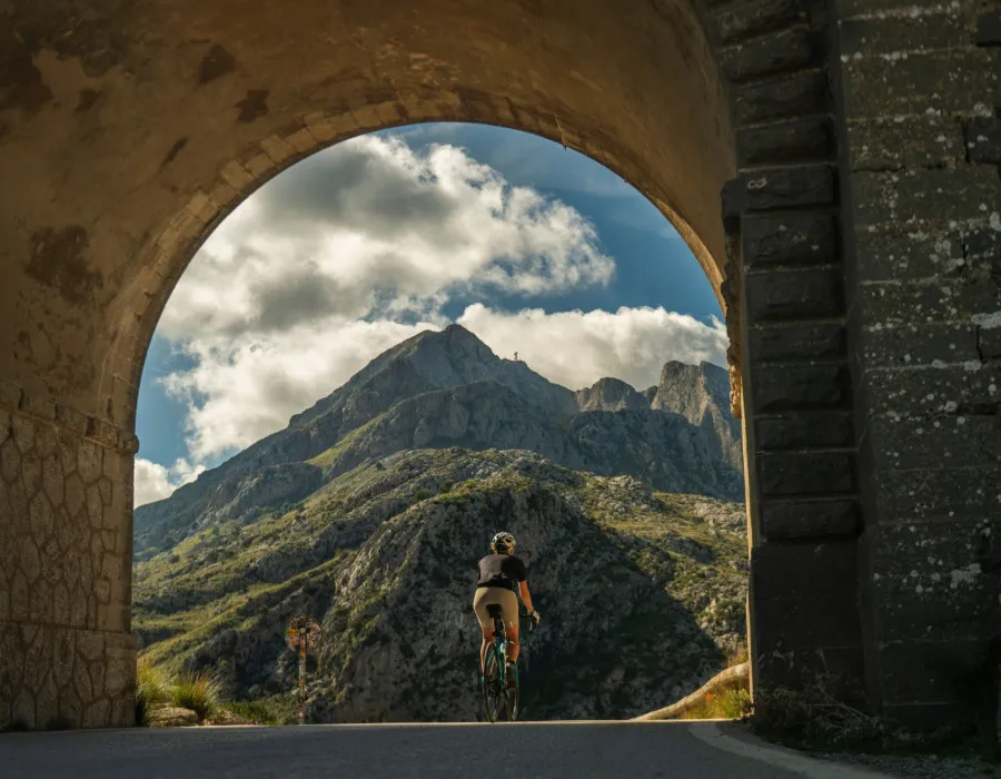 A cyclist pauses to look out across a mountainous landscape beneath a blue sky filled with scattered clouds