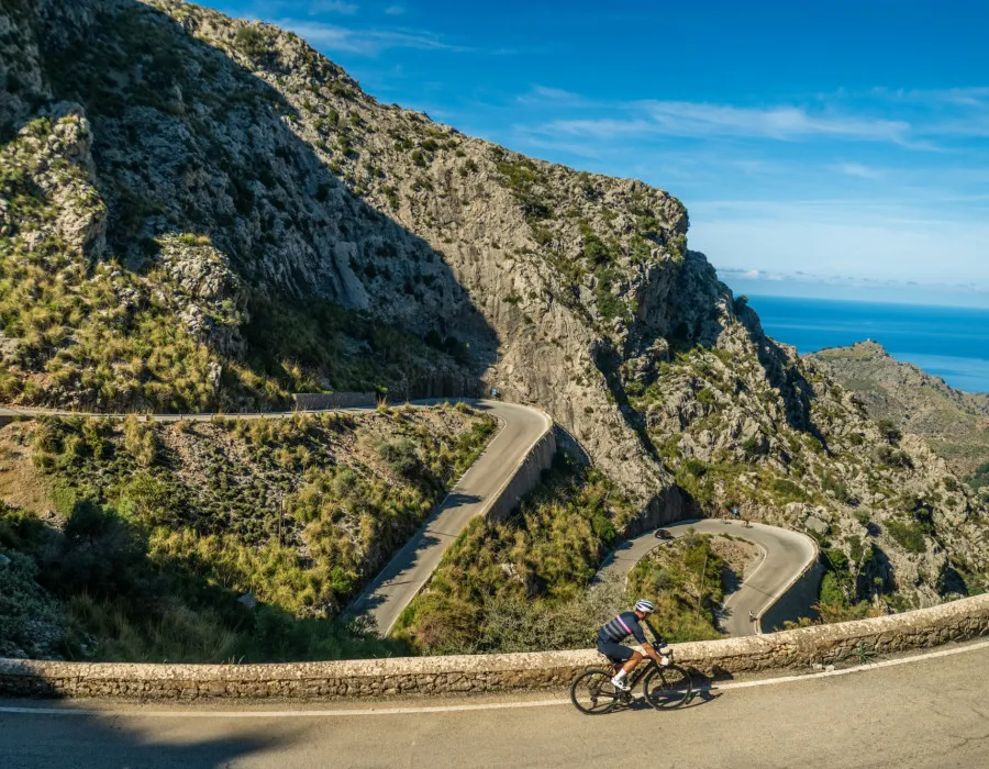 A lone cyclist rides along a winding road, with mountainous terrain, the sea, and a bright blue sky visible in the distance
