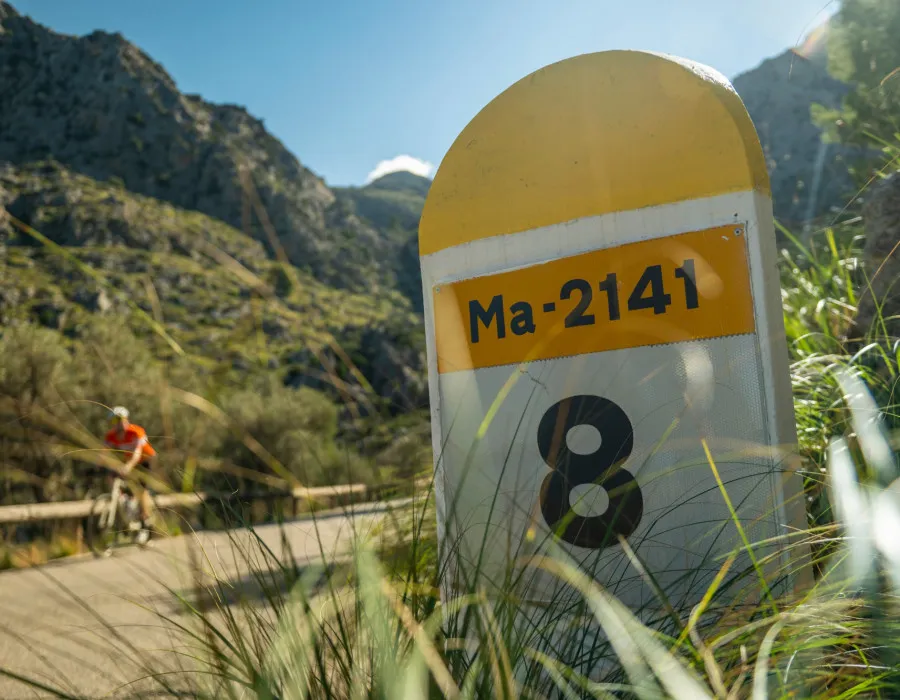 A roadside marker stands beside a winding mountain road, with a cyclist riding past in the background among rocky hills and sunlit greenery