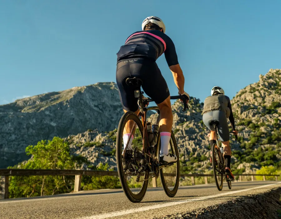 Two cyclists ride towards a mountainous landscape dotted with patches of greenery beneath a bright, sunny blue sky