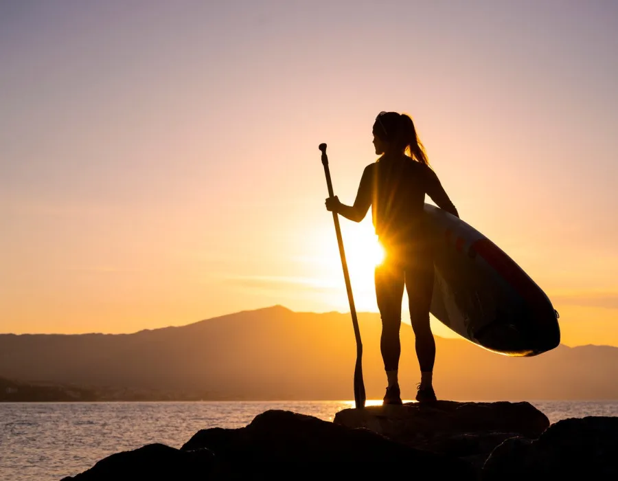 A woman stands with a paddle board, looking onto water and a pink and orange sunlit sky