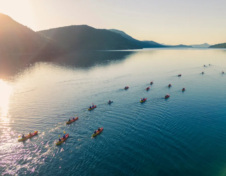 Group of participants kayaking across sunlit open water with mountains either side
