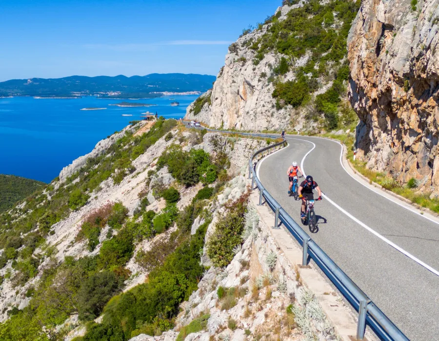 Two cyclists ride along a roadside bordered by rugged, rocky mountain terrain