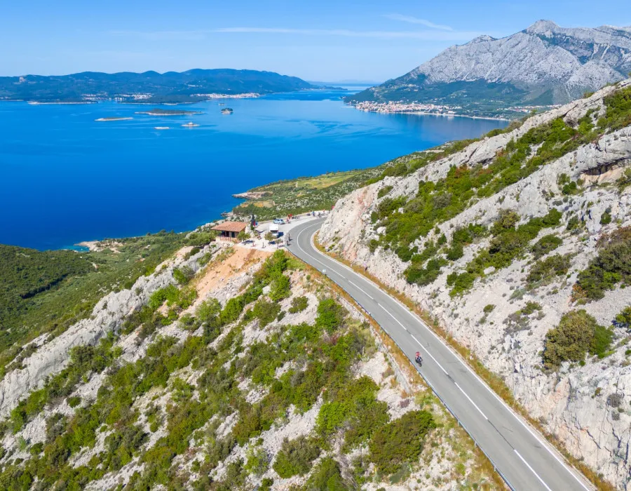 Cyclists ride along a road carved through a cliffside, with the bright blue sea visible in the background
