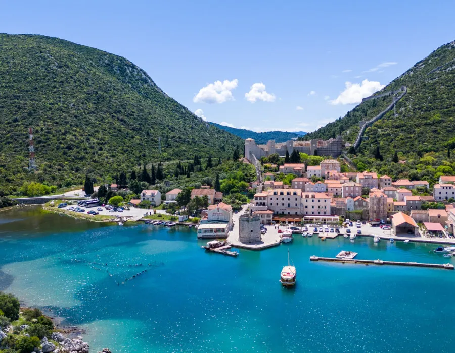 Turquoise blue water stretches towards lush green hills, with boats and waterfront buildings lining the shore in the background